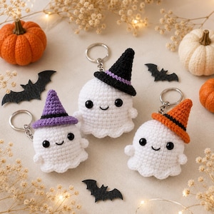 May include: Three white crocheted ghost keychains, each featuring black safety eyes and a small stitched smile. One ghost wears a purple witch hat, one wears a black witch hat with a purple band, and one wears an orange witch hat with a black band.