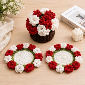 May include: A crocheted floral arrangement in a brown basket, with red and white flowers. Two matching crocheted coasters with red and white flower accents and a green border are also shown. The items are on a light wood surface.