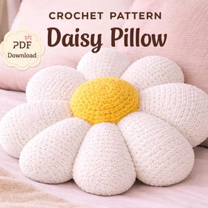 Könnte beinhalten: Gehäkeltes Gänseblümchen-Kissen mit gelber Mitte und weißen Blütenblättern. Das Kissen liegt auf einem Bett mit rosa Kissen im Hintergrund. Der Text "CROCHET PATTERN Daisy Pillow" ist oben zu sehen.