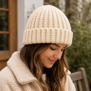 May include: A cream-colored knit beanie with a thick, folded brim and a vertical ribbed texture. The hat is constructed from a soft yarn material, featuring a rounded crown and a consistent, uniform stitch pattern throughout the entire design.