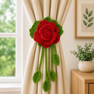 May include: A crochet curtain tieback featuring a large, vibrant red rose with layered petals and green leaves. The floral centerpiece is attached to four dangling green stems, each ending in a small, crocheted leaf, securing a beige curtain.
