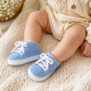 May include: A pair of light blue and white crochet baby booties designed to resemble sneakers. The booties feature a textured light blue upper, a white sole, and white crochet laces tied in a bow at the front. The construction is entirely yarn-based.