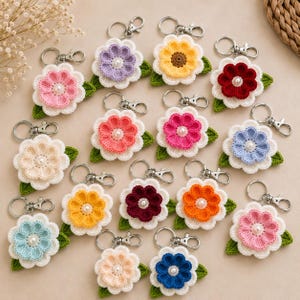 May include: A selection of crochet flower keyrings in a variety of colours, such as pink, yellow, blue, and red. Each flower features a pearl centre and a green leaf backing, attached to a silver keyring.