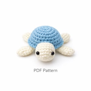 May include: A crocheted turtle with a light blue shell, cream-colored head and limbs, and black eyes. The text "PDF Pattern" is below the turtle. This handmade item is a charming, soft toy.