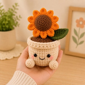 Pode incluir: Um girassol de crochê em um vaso sorridente. O girassol tem pétalas laranja e um centro marrom. O vaso é bege com um rosto sorridente, bochechas rosadas e pés bege pequenos. Uma folha verde está ao lado do girassol.
