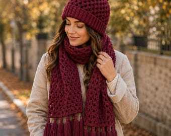 May include: A deep burgundy crochet beanie with a matching pom-pom and a long, thick crochet scarf featuring a textured open-stitch pattern and multiple dangling tassels at both ends, worn over a light beige coat in a cold outdoor setting.