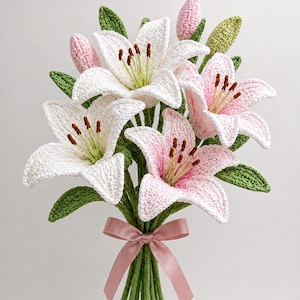 Könnte beinhalten: Ein handgefertigter Strauß gehäkelter Lilien. Der Strauß zeigt weiße und rosa Blüten mit grünen Blättern und Stielen. Ein rosa Band ist um die Stiele gebunden. Die Häkelarbeit ist detailliert und realistisch.