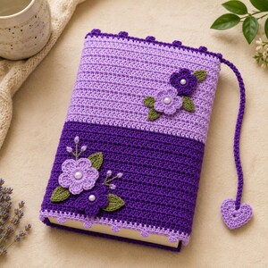 Puede incluir: Una funda de libro rectangular hecha de hilo de ganchillo en dos tonos de morado, con una sección superior morado claro y una inferior morado oscuro. La funda está decorada con flores de ganchillo en morado y lila con hojas verdes.