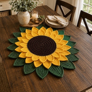 May include: A round crochet placemat designed to resemble a sunflower, featuring a dark brown center, two layers of bright yellow petals, and an outer ring of dark green leaves. The textured, handcrafted textile piece lies flat on a wooden surface.