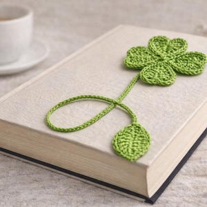 May include: A green crocheted four-leaf clover bookmark resting on a closed book. The bookmark has a stem and a leaf at the bottom. A white teacup and saucer are in the background.