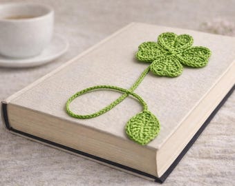May include: A green crocheted four-leaf clover bookmark resting on a closed book. The bookmark has a stem and a leaf at the bottom. A white teacup and saucer are in the background.