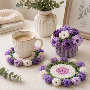 May include: A beige ceramic mug with coffee sits on a handmade crochet coaster. The coaster features a floral design with purple, white, and green accents. A matching crochet flower basket and additional coasters are also visible.