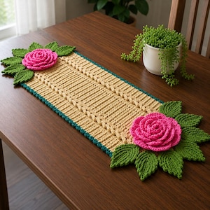 Puede incluir: Un camino de mesa rectangular de ganchillo en tono beige claro con un borde verde azulado. Cada extremo presenta una gran rosa rosa tridimensional rodeada de hojas verdes texturizadas. El centro tiene un patrón acanalado con trenzas.