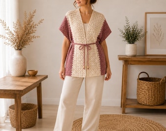 May include: A crochet cardigan featuring a cream-colored center panel and mauve-colored side panels. The garment has short sleeves, an open front, and a thin mauve tie belt at the waist with small wooden beads at the ends, paired with cream trousers.
