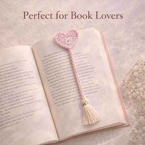 Puede incluir: Un marcapáginas de corazón rosa tejido a ganchillo con una borla, colocado en un libro abierto. El marcapáginas tiene una parte superior en forma de corazón y un tallo largo y trenzado. La borla es de color crema claro. El texto "Perfect for Book Lovers" está en la parte superior de la imagen.