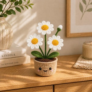 Peut inclure: Un arrangement de fleurs de marguerites crochetées dans un petit pot. Les marguerites ont des pétales blancs et des centres jaunes. Le pot est beige avec un visage souriant. Des feuilles et des tiges vertes soutiennent les fleurs.