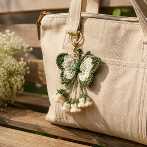 May include: A beige canvas tote bag featuring a crochet butterfly keychain. The butterfly is crafted in dark green, light green, and cream yarn, with four small cream-colored crochet bell-shaped flowers hanging from its base by thin green cords.
