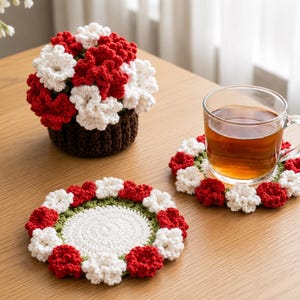 Könnte beinhalten: Ein gehäkeltes Blumenarrangement in einem braunen Topf mit roten und weißen Blumen. Zwei passende Untersetzer mit roten, weißen und grünen Blumenmustern umgeben eine Glas-Tasse mit Tee.