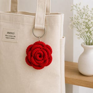 Puede incluir: Un llavero de rosa roja, estilo cuerda, cuelga de un anillo plateado sujeto a un bolso tote color crema. El bolso tiene una pequeña etiqueta que dice "merci" con texto en francés. Un pequeño jarrón blanco con flores blancas en el fondo.