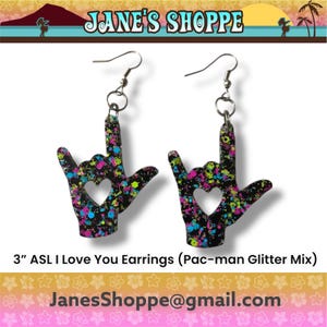 May include: A pair of earrings in the shape of the ASL "I Love You" hand sign. Each black earring has a heart cutout and is decorated with a colorful Pac-man glitter mix. The earrings are 3 inches in length.