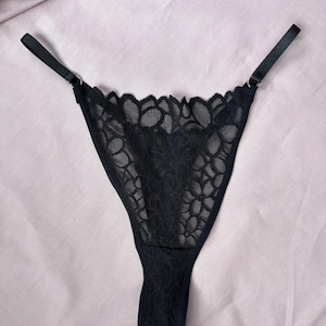May include: Black lace thong underwear with adjustable straps. The delicate floral lace design covers the front and back of the garment. The straps are thin and black, attached to the underwear with small metal rings. The underwear is displayed on a pink fabric.