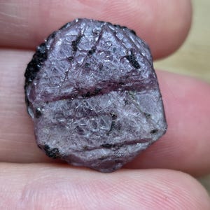 May include: A raw, translucent purple gemstone with a hexagonal shape, featuring a textured surface with visible internal inclusions, natural cracks, and small dark mineral deposits embedded within the crystalline structure of the stone.