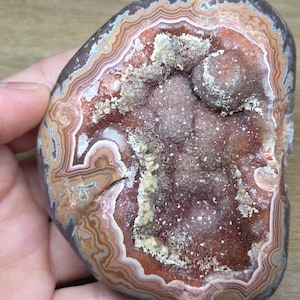May include: A polished agate stone featuring concentric bands of brown, tan, and cream colors surrounding a central cavity filled with sparkling, deep reddish-brown druzy crystals and small, textured mineral deposits throughout the center.