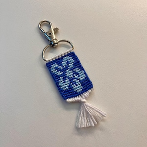 May include: A handmade blue and white keychain with a floral pattern. The rectangular woven design features light blue flowers on a dark blue background. It has a white tassel and a silver-coloured clasp.