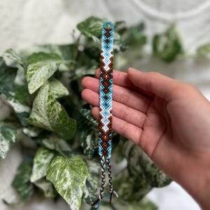 May include: Handmade friendship bracelet with a geometric design in brown, turquoise, and white. The woven threads form a pattern. It has a loop at one end and braided ties at the other.