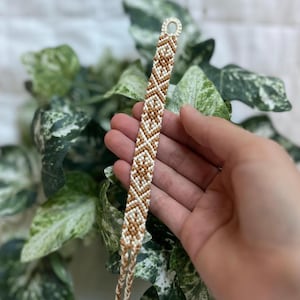 May include: A woven friendship bracelet with a geometric design in brown and white. The bracelet is made of threads and has a loop closure. The background features green and white foliage.