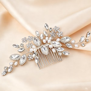 Könnte beinhalten: Ein silberner Brauthaarkamm mit floralem Design. Der Kamm hat klare, funkelnde Kristalle, Kunstperlen und kleine Blumenakzente. Der Kamm ist vor einem cremefarbenen Satinhintergrund platziert.
