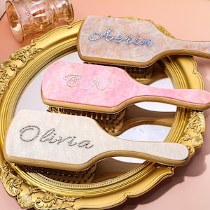 Könnte beinhalten: Drei personalisierte Haarbürsten mit Namen, die mit Strasssteinen geschrieben sind. Die Bürsten haben ein perlmuttartiges Finish in Rosa, Creme und Pfirsich, mit goldfarbenen Holzgriffen. Sie werden auf einem goldgerahmten Spiegel ausgestellt.