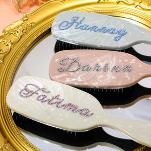 Könnte beinhalten: Drei personalisierte Haarbürsten mit Namen, die mit Strasssteinen geschrieben sind. Die Bürsten sind in Pastellfarben: Hellgrau, Rosa und Off-White. Die Namen sind "Hannay", "Darina" und "Fatima". Die Bürsten liegen auf einer reflektierenden Oberfläche.