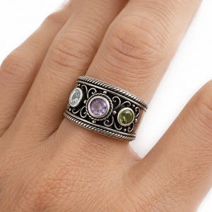 May include: A wide silver band ring featuring three round bezel-set gemstones in a row. The center stone is purple, flanked by a light blue stone on the left and a green stone on the right. The silver band has intricate scrollwork and rope edges.