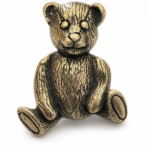 May include: A small, seated teddy bear figurine crafted from a metallic material with an antique brass finish. The surface features a textured, fur-like pattern throughout, with defined ears, a rounded snout, and oval-shaped feet extending outward.