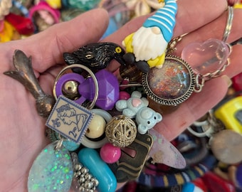 May include: A selection of colourful charms and trinkets, including a gnome, a black bird, and various beads and pendants. The items are held in a hand, with a background of other assorted objects.