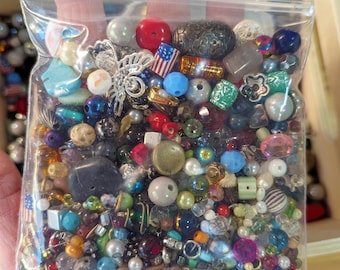 May include: A clear plastic bag filled with a variety of colourful beads, including glass, metal, and plastic. The beads are in various shapes and sizes, and some have a metallic finish. There are also a few small charms, including a butterfly and a star.
