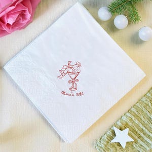 May include: White square paper napkin with a red illustration of a person relaxing in a martini glass. The text "Olivia's 30th" is printed below the illustration. The napkin is surrounded by a pink rose, white ornaments, and a star.
