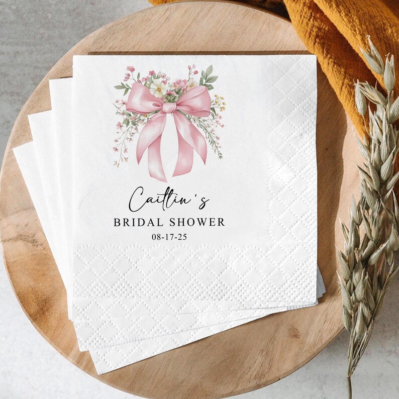 May include: White paper napkins with a pink bow and floral design, with the text "Caitlin's Bridal Shower" and the date 08-17-25. The napkins are stacked on a wooden tray.