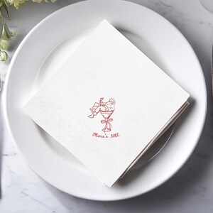 May include: White cocktail napkins with a red illustration of a person in a martini glass, with the text "Olivia's 30th". The napkins are folded and placed on a white plate, suitable for a birthday celebration.