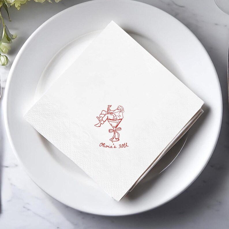 May include: White cocktail napkins with a red illustration of a person in a martini glass, with the text "Olivia's 30th". The napkins are folded and placed on a white plate, suitable for a birthday celebration.