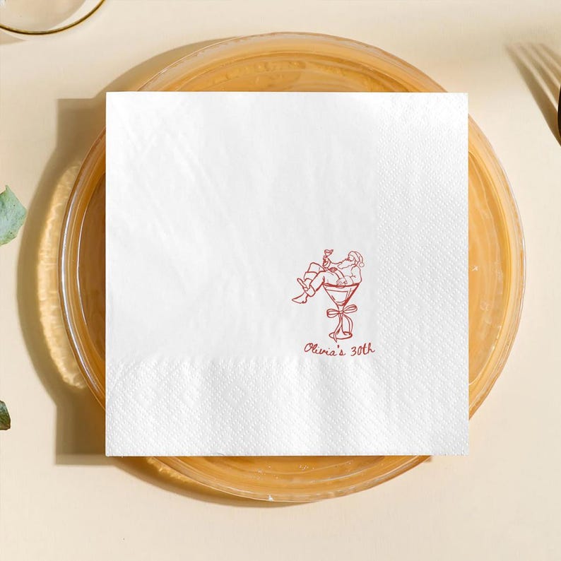 May include: White square paper napkin with a red illustration of a person relaxing in a martini glass. The text "Olivia's 30th" is written in red script below the illustration. The napkin is on a clear plate.