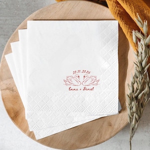 May include: White paper napkins with a diamond pattern. The napkins feature a red illustration of two swans facing each other, with the date 20.11.2025 and the names Emma + Daniel printed below. The napkins are stacked on a wooden plate.