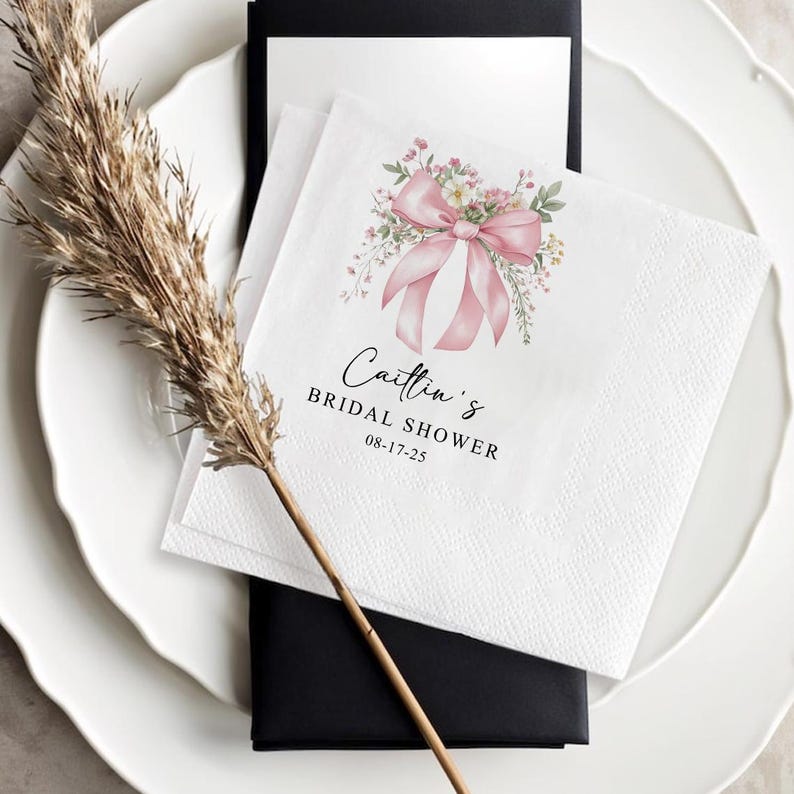 May include: White paper napkins with a pink bow and floral design, with the text "Caitlin's Bridal Shower" and the date 08-17-25. The napkins are placed on a white plate with a black card underneath.