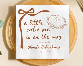 May include: A white square napkin with brown text and a pie illustration. The text reads "a little cutie pie is on the way" and "Olivia's Baby shower" with a date. A brown ribbon design is at the top.