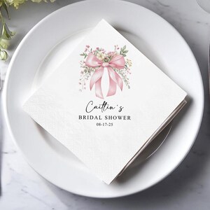 May include: A white paper napkin with a pink bow and floral design, with the text "Caitlin's BRIDAL SHOWER 08-17-25". The napkin is folded and placed on a white plate, suitable for a bridal shower.