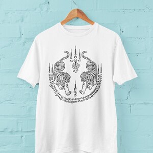 May include: White t-shirt with a black illustration of two tigers facing each other, encircled by text and vertical lines. The tigers have detailed stripes and are symmetrically placed. The shirt hangs on a wooden hanger against a light blue wall.