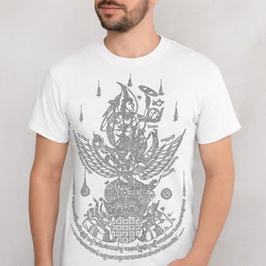 Puede incluir: Camiseta blanca con una detallada ilustración gris de una figura mitológica con alas, patrones intrincados y texto. El diseño está centrado en la camiseta. La camiseta tiene mangas cortas y cuello redondo.