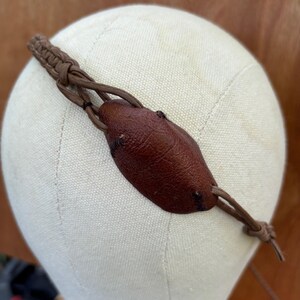 May include: A brown leather eye patch with a braided headband and adjustable ties. The eye patch is oval-shaped and has a textured surface. The headband and ties are made of a lighter brown material.