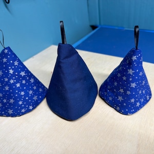 May include: Three blue fabric items with a conical shape and a small black loop at the top. Two are patterned with silver stars, while the center one is solid blue. They are arranged on a light wood surface.